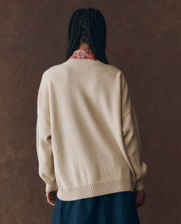 A person with braided hair, seen from behind, wears THE GREAT. F25 D1’s The Stable Pullover in Natural and a red patterned scarf against a brown textured background.