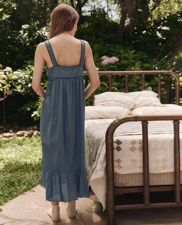 A woman wearing THE GREAT. SP25 SLEEP's The Western Tank Night Dress in Blue Dusk stands with her back to the camera beside a vintage brass bed in a garden.