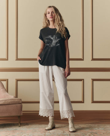 A woman stands indoors against a beige paneled wall, wearing THE GREAT. SP26 D2 The Boxy Crew W/ Shadow Oak Graphic in washed black, white wide-leg pants with lace hems, and light-colored lace-up boots.