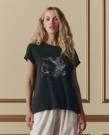 A person with long, wavy blonde hair wears THE GREAT.'s The Boxy Crew W/ Shadow Oak Graphic in washed black and white pants, standing by a beige wall with decorative molding.