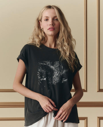 A woman with long blonde hair wears THE GREAT.’s The Boxy Crew W/ Shadow Oak Graphic in washed black, styled with white pants, standing before a beige wall featuring decorative trim and shadow oak accents.