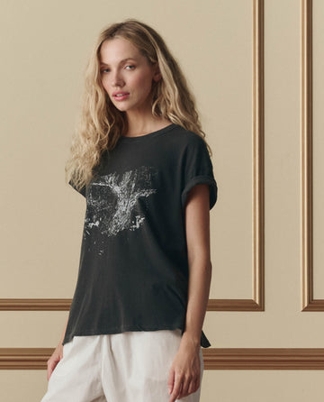 A woman with long blonde hair wears THE GREAT.'s The Boxy Crew W/ Shadow Oak Graphic in washed black and white pants, standing in front of a beige wall with decorative molding.