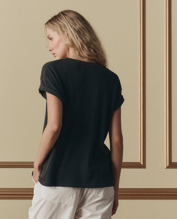 A woman with long blonde hair stands facing away, wearing THE GREAT. SP26 D2 The Boxy Crew W/ Shadow Oak Graphic in washed black and white pants against a beige wall with decorative molding.