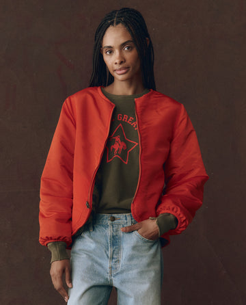 A person stands against a brown background wearing THE GREAT. F25 D1's The Reversible Plush Bomber in Camo & Hot Red over a green sweatshirt with a star and horse graphic, paired with light blue jeans.