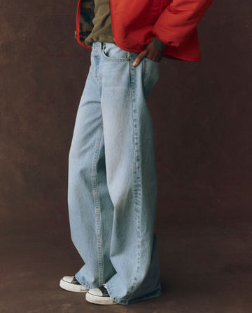 A person wears The Slouch Wide Leg Jean in Tidal Wash by THE GREAT. F25 D1, paired with a red jacket and black-and-white sneakers, standing against a dark brown background.