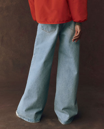 A person wears THE GREAT. F25 D1 The Slouch Wide Leg Jean — Tidal Wash, featuring classic five-pocket construction, with white shoes and a red jacket, standing on a dark background, back partially turned.