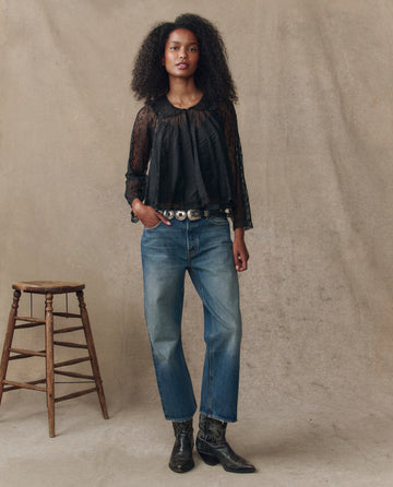 A woman stands against a neutral backdrop wearing THE GREAT. H25 COLLECTION's Keepsake Lace Collared Top in black, paired with blue jeans, a silver-buckled belt, black boots, and a wooden stool to her left.