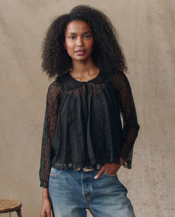 A woman with curly hair models THE GREAT. H25 COLLECTION’s The Keepsake Lace Collared Top in black, paired with blue jeans, against a beige backdrop with a wooden stool beside her.