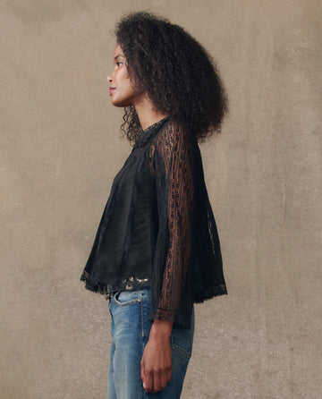 A person with curly hair stands in profile against a plain background, wearing THE GREAT. H25 COLLECTION's The Keepsake Lace Collared Top in black and blue jeans.