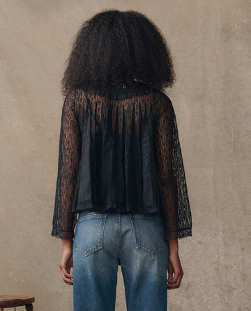 A person stands with their back to the camera, wearing THE GREAT. H25 COLLECTION’s The Keepsake Lace Collared Top in black and blue jeans against a neutral background.