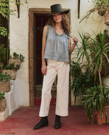 A person in THE GREAT. PF25 The Vignette Top in Pale Blue, white pants, black boots, and a wide-brimmed black hat stands indoors among potted plants.