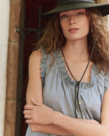 A woman with wavy hair, wearing a wide-brimmed black hat and the Pale Blue Vignette Top by THE GREAT. PF25, stands in front of a doorway with her arms crossed.
