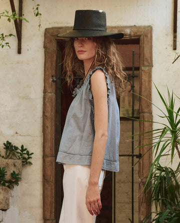 A woman with curly hair wears a wide-brimmed black hat, a white skirt, and THE GREAT. PF25 The Vignette Top in pale blue as she stands sideways in a rustic outdoor setting surrounded by plants.