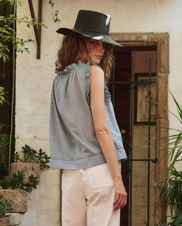 Wearing THE GREAT. PF25's The Vignette Top in Pale Blue, a person with a wide-brimmed black hat and light pants stands outdoors by a stone wall and doorway, glancing over their shoulder.