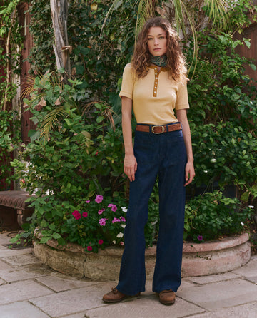 A woman stands outdoors on a stone patio, surrounded by greenery. She wears THE GREAT. PF25's The Baseline Henley in Bronze Stripe, blue flared jeans, a brown belt, and brown shoes.