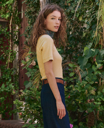 A woman with wavy brown hair stands sideways outdoors amid green foliage, wearing THE GREAT. PF25’s The Baseline Henley in Bronze Stripe, dark blue pants, and a patterned scarf around her neck.