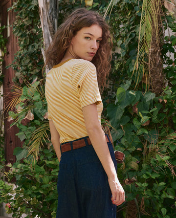 A woman with curly hair stands outdoors among green plants, looking over her shoulder while wearing THE GREAT. PF25 The Baseline Henley in Bronze Stripe and blue jeans.