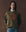 A person wears THE GREAT. F25 D1 The College Sweatshirt in Dark Army with a Rodeo Star graphic, standing against a brown backdrop with hands in pockets.