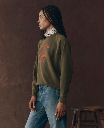 A person with braided hair wears THE GREAT. F25 D1's The College Sweatshirt in Dark Army with Rodeo Star Graphic, paired with blue jeans and a white patterned scarf, standing beside a wooden stool against a brown background.