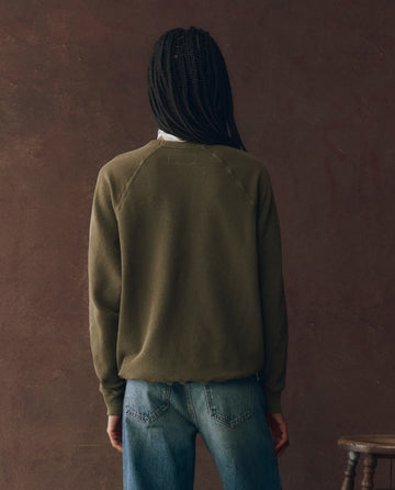 A person with braided hair stands facing away, wearing THE GREAT. F25 D1 "The College Sweatshirt" in Dark Army with Rodeo Star Graphic and blue jeans against a brown textured background.