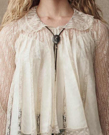 Person wearing The Keepsake Lace Collared Top in cream from THE GREAT. H25 COLLECTION, featuring sheer, lacy fabric with vertical stripes, a lace collar, and a decorative bolo tie with a silver pendant.