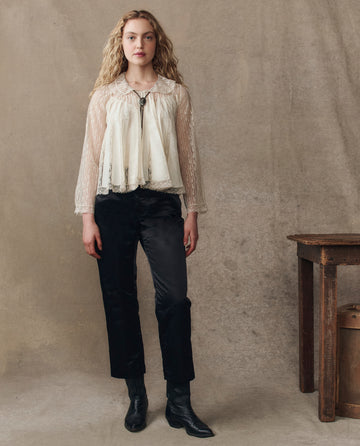 A person stands indoors beside a rustic wooden table, wearing THE GREAT. H25 COLLECTION’s The Keepsake Lace Collared Top in Cream, styled with black pants and boots, set against a neutral textured backdrop.