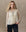 A woman with long wavy blonde hair wears THE GREAT. H25 COLLECTION's The Keepsake Lace Collared Top in cream, styled with a bolo tie and black pants, standing against a neutral textured background.