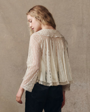 A person with long blonde hair models THE GREAT. H25 COLLECTION’s The Keepsake Lace Collared Top in cream, styled with black pants, standing against a neutral background and looking to the side.