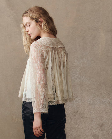 A person with wavy blonde hair wears THE GREAT. H25 COLLECTION The Keepsake Lace Collared Top in cream and black pants, standing side and back to the camera against a neutral background.