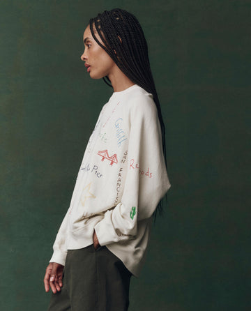 Woman with long braids wears THE GREAT.'s The Slouch Sweatshirt—Embroidered, in washed white with Golden State embroidery—styled with green pants, standing in profile against a dark green background.