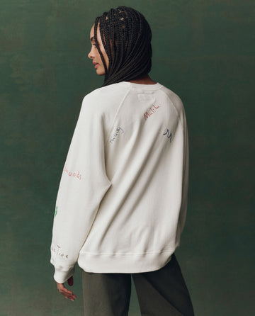 A person with braided hair wears THE GREAT. F25 D2 The Slouch Sweatshirt in Washed White with Golden State embroidery on the back, posing against a green background.