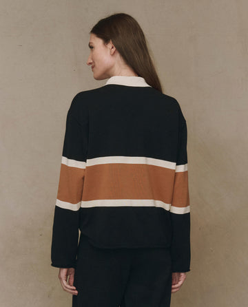 A person faces away in THE GREAT. F25 HIGH TWIST The Rugby Sweater, Novelty—black with bronze stripes, featuring dropped shoulders, paired with black pants, against a plain beige background.