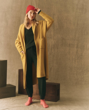A person in a yellow coat, black outfit, and red socks stands indoors against a neutral backdrop while wearing The Cashmere Hat in Bright Red by THE GREAT. HOL 22 D2 CASHMERE.