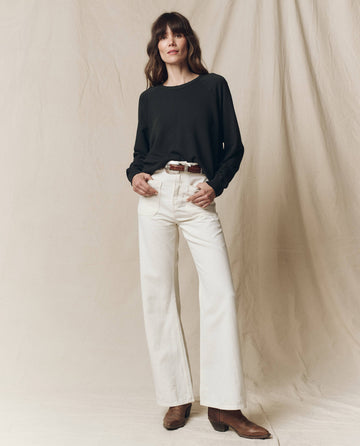 Against a beige backdrop, a person wears THE GREAT. CORE KNITS' The College Sweatshirt in Almost Black with white wide-leg jeans, a brown belt, and boots. Hands are casually tucked in pockets.