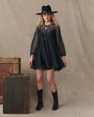 A woman models THE GREAT. H25 COLLECTION's The Keepsake Lace Shift Dress in black, paired with boots and a hat, standing indoors by wooden crates on a beige backdrop, capturing a chic, memorable moment.