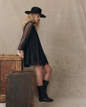 A woman with long blonde hair wears THE GREAT. H25 COLLECTION’s The Keepsake Lace Shift Dress in black, styled with a wide-brim hat and boots, standing beside rustic wooden boxes against a neutral background.