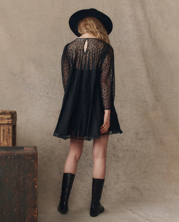 Someone wearing THE GREAT. H25 COLLECTION’s The Keepsake Lace Shift Dress in black faces away from the camera, styled with a black hat and boots, against a neutral backdrop with wooden crates nearby.