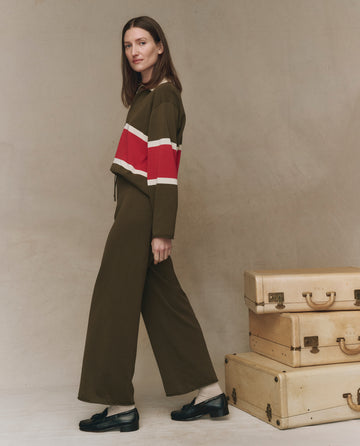 A woman in THE GREAT. F25 HIGH TWIST The Rec Sweater Pant in Army walks past two stacked vintage suitcases against a neutral background.