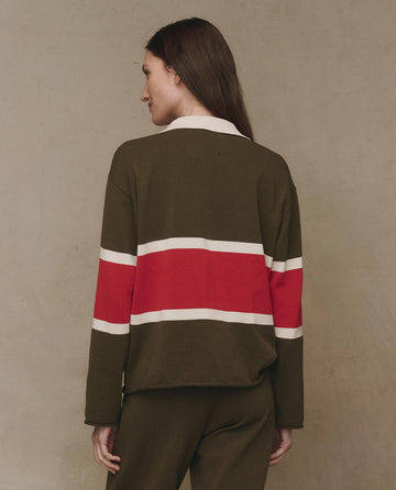 A woman with long brown hair is seen from behind wearing THE GREAT. F25 HIGH TWIST’s Rugby Sweater in Army with Hot Red Stripe, paired with coordinating pants, pictured against a neutral background.
