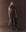 A woman stands against a textured brown background wearing THE GREAT. F25 D1's The Tulsa Dress in Geo Bandana Print, styled with a black necktie and dark cowboy boots.