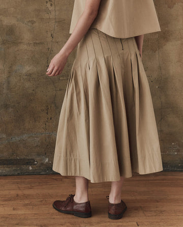 Someone wears THE GREAT. SP25 POPLIN The Rhythm Skirt in Khaki with a matching top and brown oxfords, standing on a wooden floor against a textured brown wall for a timeless style.