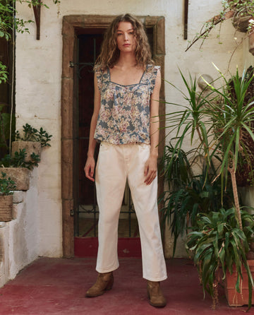 A woman stands indoors on a red floor, wearing THE GREAT. PF25's The Victorian Pleat Tank in Indigo Floral, paired with white pants and brown boots, surrounded by green plants against a white brick wall.