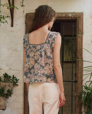 A person with wavy brown hair wears THE GREAT. PF25’s The Victorian Pleat Tank in Indigo Floral and white pants, standing with their back to a rustic white wall and doorway, with plants nearby.