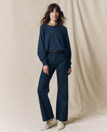 A person wearing The College Sweatshirt in True Navy from THE GREAT. FALL 23 KNITS, paired with high-waisted dark jeans and white sneakers, stands against a light fabric backdrop for a relaxed casual vibe.