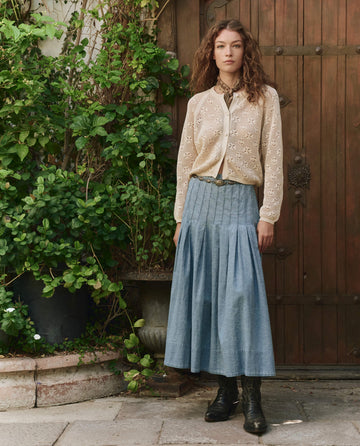 A woman stands in front of a wooden door, wearing a cream lace blouse, a pleated blue skirt, black boots, and a belt, with greenery and potted plants beside her.