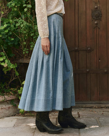 A person stands outdoors wearing a long light blue denim skirt, black boots, and a cream-colored lace blouse, with plants and a wooden door in the background.