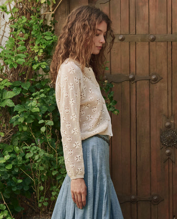A woman with curly hair stands in profile wearing THE GREAT. PF25 The Crochet Cardigan in Natural and a blue skirt, beside a wooden door and green foliage.