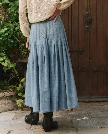 A person wearing a cream lace long-sleeve top and a pleated blue denim maxi skirt stands on stone pavement in front of a wooden door, facing away from the camera.