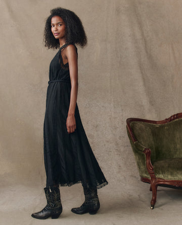A woman wearing THE GREAT. H25 COLLECTION’s The Keepsake Lace Column Dress in black and black cowboy boots stands beside a vintage green armchair against a beige backdrop.