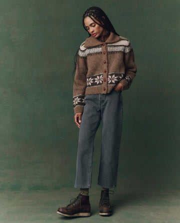 A person stands against a green background, wearing THE GREAT. F25 D2’s The Wayne Jean in Graphite Wash with Clean Hem, paired with a brown patterned sweater and boots, one hand in their pocket, looking down.
